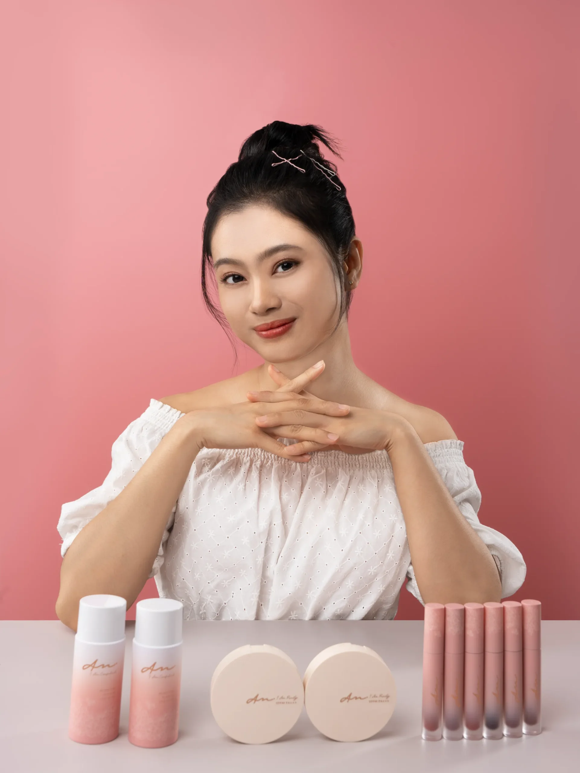 Confident woman with an elegant updo hairstyle, wearing a white off-the-shoulder blouse, poses behind a display of 'Alyssa Madeline' beauty products, including two bottles, two compact powders, and five lip glosses, all arrayed neatly against a soft pink background.