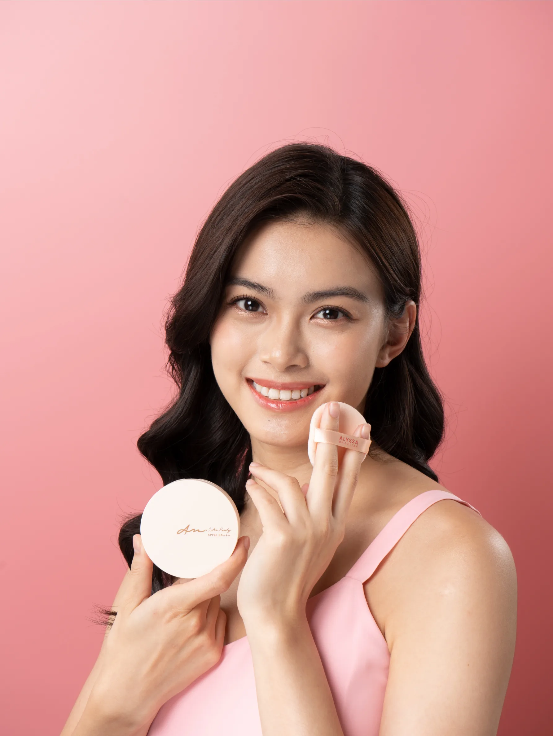 Confident woman with an elegant updo hairstyle, wearing a white off-the-shoulder blouse, poses behind a display of 'Alyssa Madeline' beauty products, including two bottles, two compact powders, and five lip glosses, all arrayed neatly against a soft pink background.